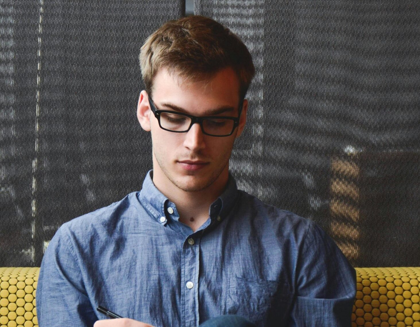 A young man in glasses writes in a notebook while sitting on a stylish couch indoors.